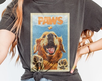 May include: A charcoal gray short-sleeve t-shirt featuring a rectangular graphic print of a golden retriever dog with its paws raised. The text PAWS is printed in orange above the dog, which is looking up at a small treat floating in the air.