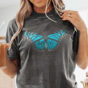 May include: A charcoal gray short-sleeve t-shirt featuring a large, vibrant blue butterfly graphic centered on the chest. The butterfly has detailed black wing veins and white spots along the edges of its wings. The shirt has a crew neckline.