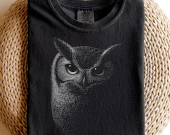 May include: A black crew neck t-shirt featuring a detailed white stippled illustration of an owl's face on the front. The owl is depicted with large, focused eyes and textured feathers, with the design fading into the black fabric of the shirt.