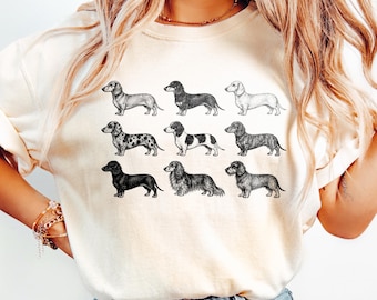 May include: A cream-colored short-sleeve t-shirt featuring a graphic print of nine dachshund dogs in various coat patterns and colors, including solid black, spotted, and long-haired varieties, arranged in three rows of three on the front.