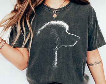 May include: A charcoal gray short-sleeve t-shirt featuring a white line-art graphic of a poodle in profile. The shirt has a crew neckline and a vintage-washed appearance. The design is centered on the chest, showcasing the dog's curly fur texture.