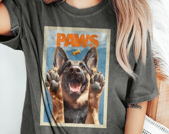 May include: A charcoal gray t-shirt featuring a rectangular graphic print of a German Shepherd dog with its front paws raised. The word PAWS is printed in orange above the dog, with a small yellow bone icon centered between the word and the dog.