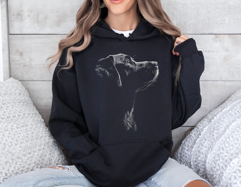 May include: A black hooded sweatshirt featuring a white line-art graphic of a dog in profile on the chest. The hoodie includes a front kangaroo pocket, long sleeves with ribbed cuffs, and a drawstring hood, providing a casual and functional garment.
