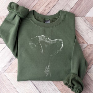 May include: An olive green crewneck sweatshirt featuring a white line art illustration of a dog in profile on the chest. The garment has long sleeves with ribbed cuffs and a ribbed neckline, showcasing a simple and clean design on the front.
