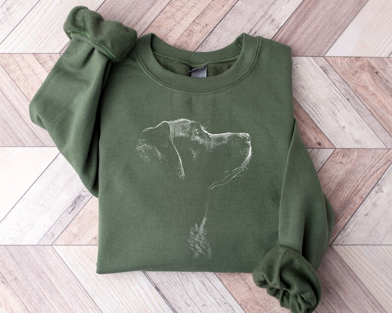 May include: An olive green crewneck sweatshirt featuring a white line art illustration of a dog in profile on the chest. The garment has long sleeves with ribbed cuffs and a ribbed neckline, showcasing a simple and clean design on the front.