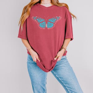 May include: A maroon short-sleeved t-shirt featuring a large, centered graphic of a butterfly with bright turquoise wings and white accents. The shirt has a crew neckline and a relaxed, loose-fitting silhouette, providing a casual appearance.