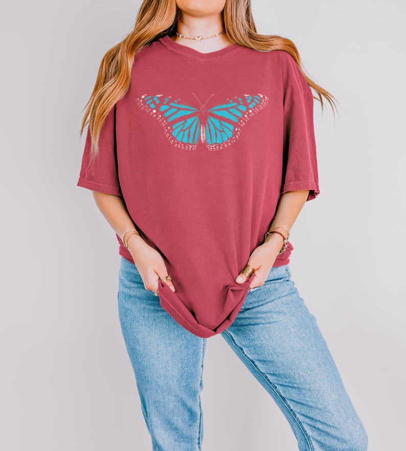 May include: A maroon short-sleeved t-shirt featuring a large, centered graphic of a butterfly with bright turquoise wings and white accents. The shirt has a crew neckline and a relaxed, loose-fitting silhouette, providing a casual appearance.