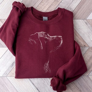 May include: A maroon crewneck sweatshirt featuring a white line-art illustration of a dog's profile on the chest. The garment has long sleeves, a ribbed neckline, and a Gildan brand label visible inside the collar, showcasing a simple canine graphic.