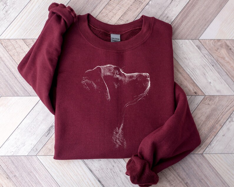 May include: A maroon crewneck sweatshirt featuring a white line-art illustration of a dog's profile on the chest. The garment has long sleeves, a ribbed neckline, and a Gildan brand label visible inside the collar, showcasing a simple canine graphic.