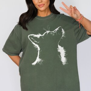May include: A dark olive green short-sleeved t-shirt featuring a white graphic illustration of a dog looking upward. The design captures the silhouette of the dog's head and neck with detailed line work, set against the solid colored fabric.