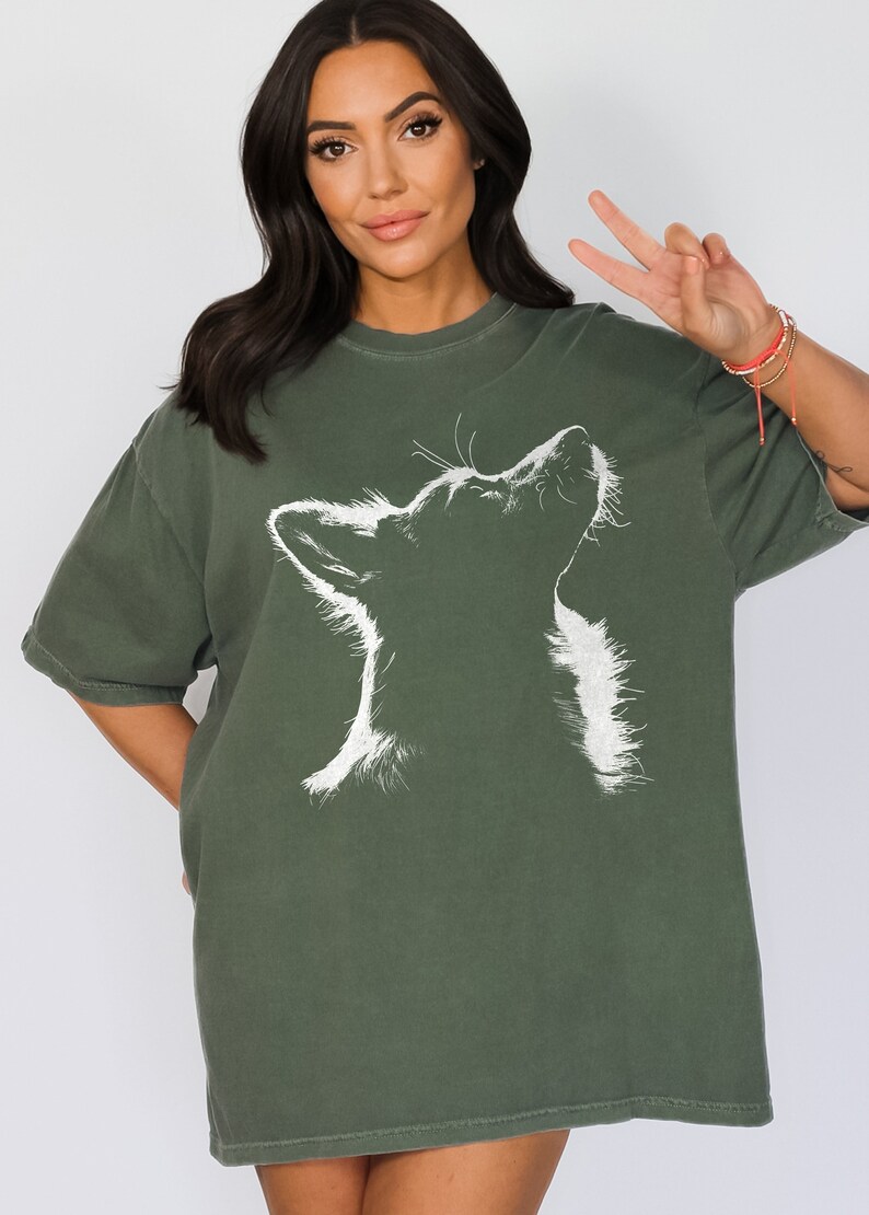 May include: A dark olive green short-sleeved t-shirt featuring a white graphic illustration of a dog looking upward. The design captures the silhouette of the dog's head and neck with detailed line work, set against the solid colored fabric.