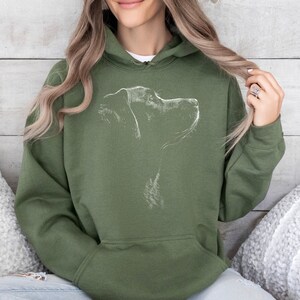 May include: An olive green hooded sweatshirt featuring a white line art illustration of a dog's profile on the chest. The garment includes a drawstring hood, long sleeves, and a large front kangaroo pocket, providing a casual and functional design.