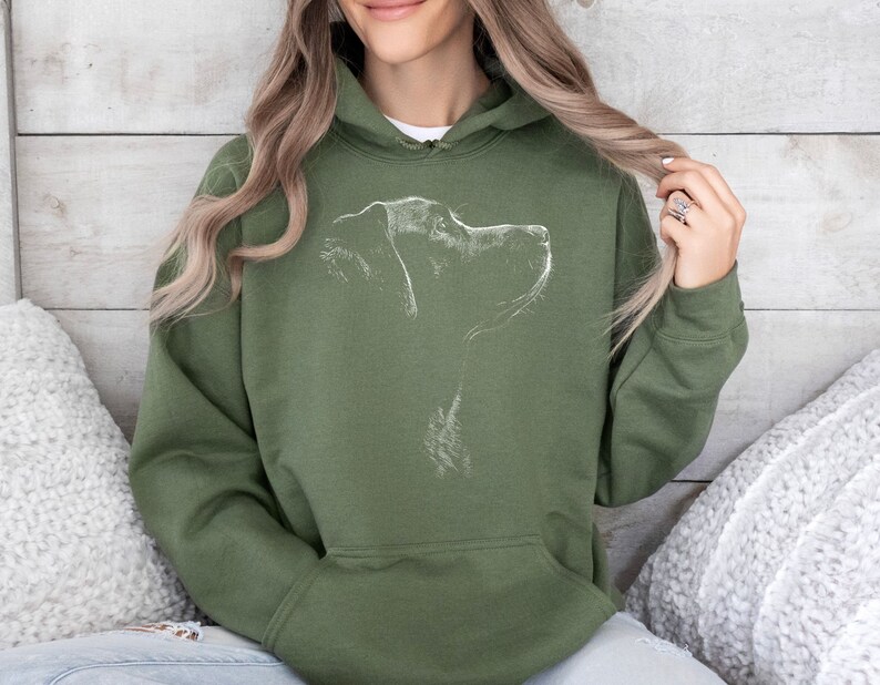 May include: An olive green hooded sweatshirt featuring a white line art illustration of a dog's profile on the chest. The garment includes a drawstring hood, long sleeves, and a large front kangaroo pocket, providing a casual and functional design.