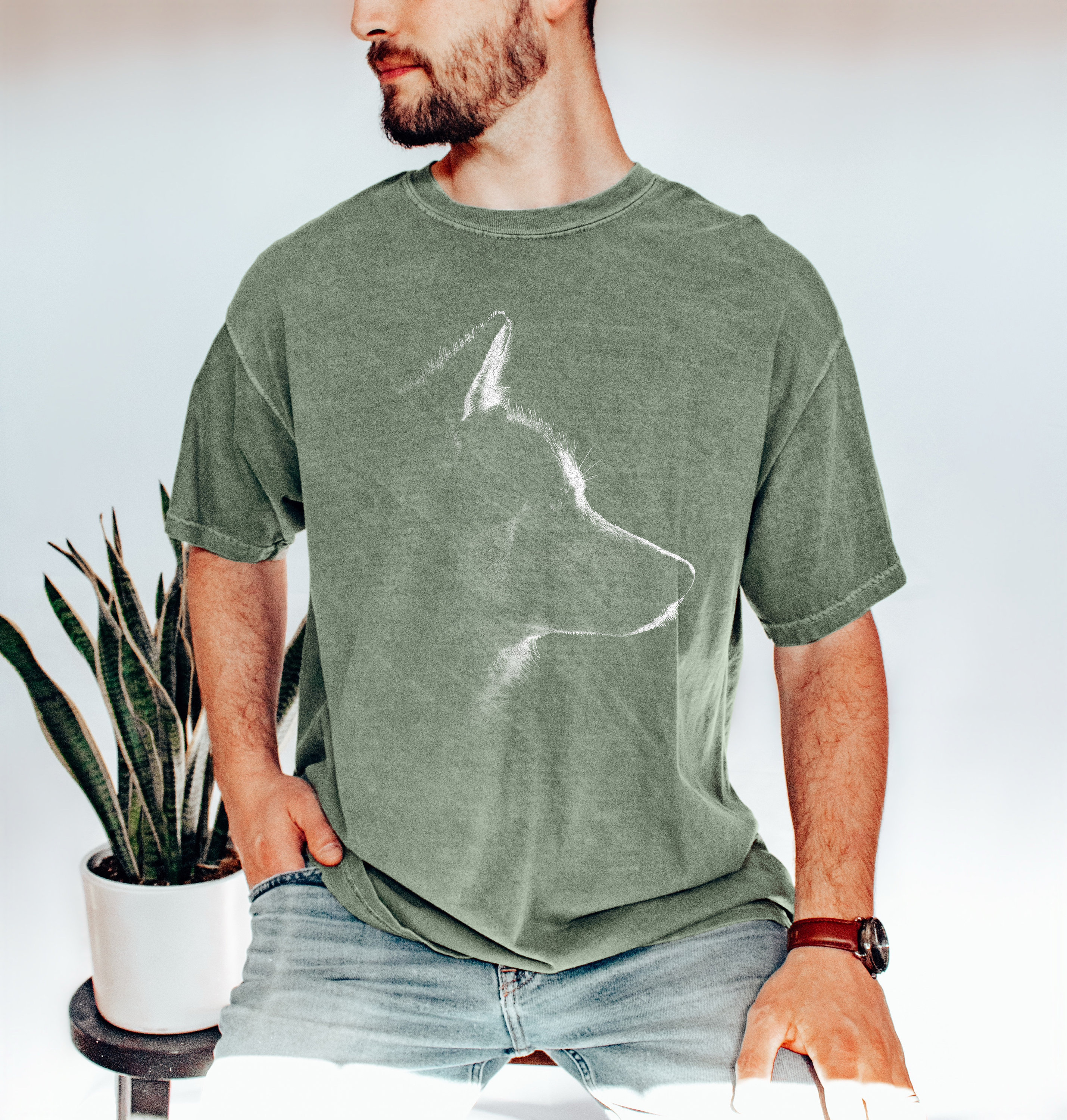 May include: A sage green short-sleeve crew neck t-shirt featuring a white line art graphic of a dog's profile silhouette centered on the chest. The fabric has a slightly faded, vintage-inspired texture and a relaxed fit with short sleeves.