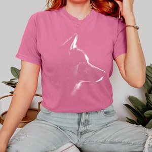 May include: A pink short-sleeved t-shirt featuring a white graphic outline of a dog's head in profile. The shirt has a crew neckline and a relaxed fit, showcasing a minimalist artistic representation of a canine silhouette centered on the chest.