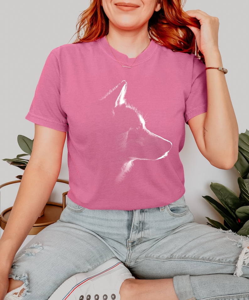 May include: A pink short-sleeved t-shirt featuring a white graphic outline of a dog's head in profile. The shirt has a crew neckline and a relaxed fit, showcasing a minimalist artistic representation of a canine silhouette centered on the chest.