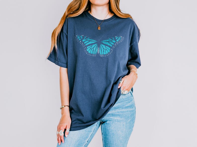 May include: A dark blue short-sleeved t-shirt featuring a large, detailed graphic of a teal butterfly centered on the chest. The butterfly design includes intricate wing patterns with white accents along the edges, set against the solid dark blue fabric.