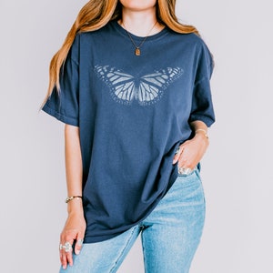 May include: A navy blue short-sleeve t-shirt featuring a large, centered graphic of a monarch butterfly in a light gray, distressed print. The shirt has a crew neckline and a relaxed, loose fit, paired with light blue denim jeans.