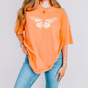 May include: An orange short-sleeved t-shirt featuring a large, white, detailed graphic of a butterfly centered on the chest. The shirt has a crew neckline and a relaxed, loose fit, paired with blue denim jeans that have a classic medium wash finish.