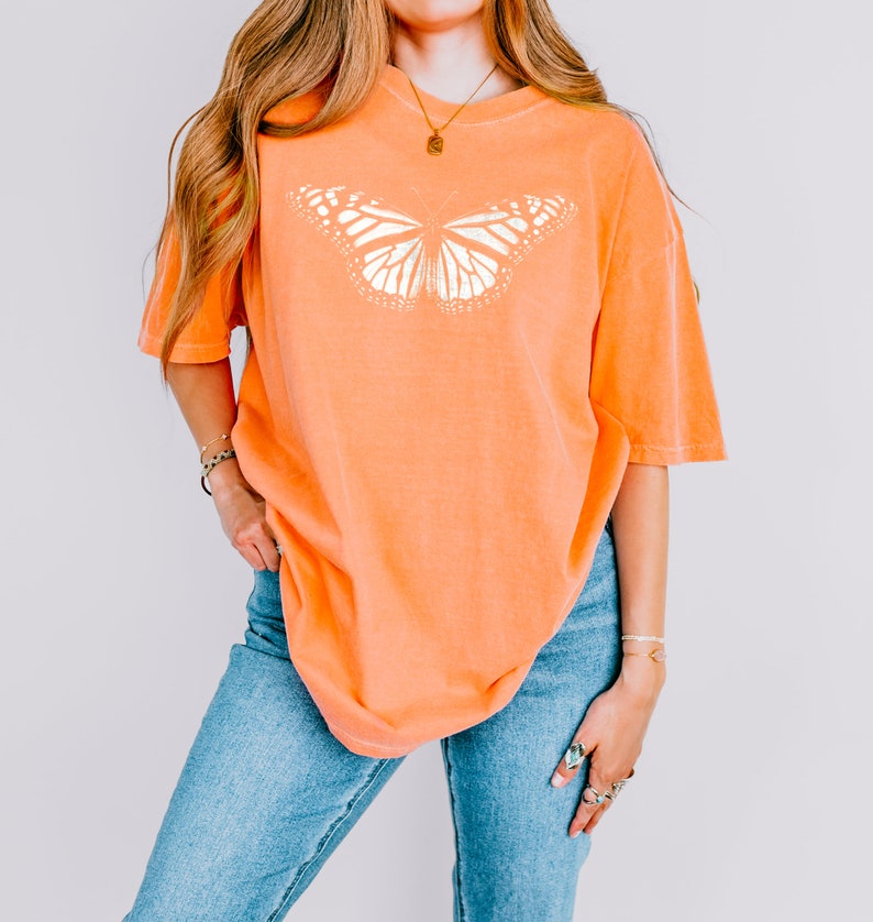 May include: An orange short-sleeved t-shirt featuring a large, white, detailed graphic of a butterfly centered on the chest. The shirt has a crew neckline and a relaxed, loose fit, paired with blue denim jeans that have a classic medium wash finish.