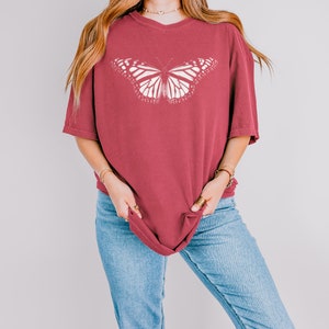 May include: A dark red, short-sleeved t-shirt featuring a large, white, detailed monarch butterfly graphic centered on the chest. The shirt has a crew neckline and a relaxed, loose-fitting silhouette, paired with light blue denim jeans.