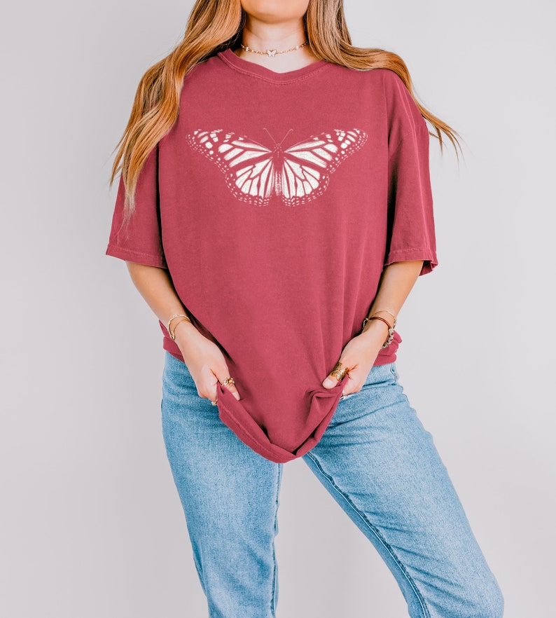 May include: A dark red, short-sleeved t-shirt featuring a large, white, detailed monarch butterfly graphic centered on the chest. The shirt has a crew neckline and a relaxed, loose-fitting silhouette, paired with light blue denim jeans.