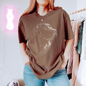 Op de afbeelding: Een bruin t-shirt met korte mouwen en een witte lijntekening van een hondenkop op de borst. Het shirt heeft een ronde hals en een relaxte pasvorm, gecombineerd met een blauwe spijkerbroek. De afbeelding toont het profiel van de hond.