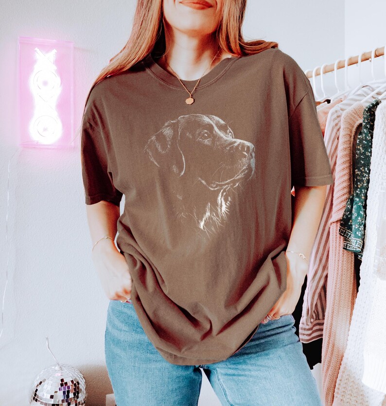 Op de afbeelding: Een bruin t-shirt met korte mouwen en een witte lijntekening van een hondenkop op de borst. Het shirt heeft een ronde hals en een relaxte pasvorm, gecombineerd met een blauwe spijkerbroek. De afbeelding toont het profiel van de hond.