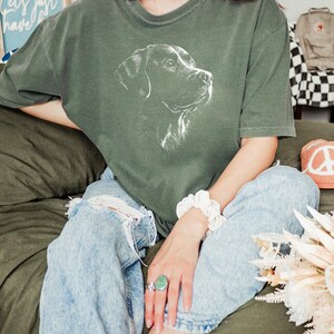 Op de afbeelding: Een donker olijfgroen T-shirt met korte mouwen en een witte lijntekening van een hondenkop op de borst. Het shirt heeft een ronde hals en een relaxte pasvorm, gecombineerd met een lichtblauwe acid-wash spijkerbroek met kniegaten.
