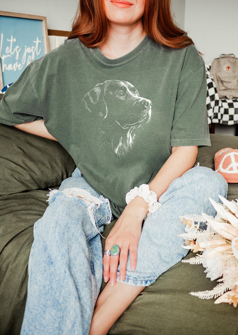 Op de afbeelding: Een donker olijfgroen T-shirt met korte mouwen en een witte lijntekening van een hondenkop op de borst. Het shirt heeft een ronde hals en een relaxte pasvorm, gecombineerd met een lichtblauwe acid-wash spijkerbroek met kniegaten.
