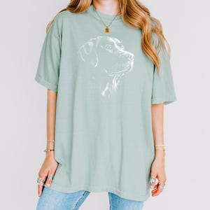 Op de afbeelding: Een saliegroen T-shirt met korte mouwen en een witte lijntekening van een hondenkop op de borst. Het shirt heeft een ronde hals en een losse pasvorm. Het ontwerp toont het profiel van een hond met gedetailleerde arceringen.