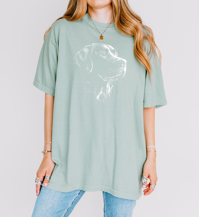 Op de afbeelding: Een saliegroen T-shirt met korte mouwen en een witte lijntekening van een hondenkop op de borst. Het shirt heeft een ronde hals en een losse pasvorm. Het ontwerp toont het profiel van een hond met gedetailleerde arceringen.