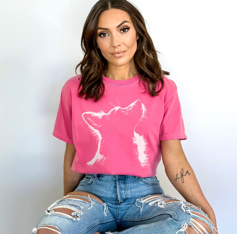 May include: A bright pink short-sleeved t-shirt featuring a white graphic outline of a dog looking upward. The shirt has a crew neckline and a relaxed fit. The design is centered on the chest, showcasing the silhouette of the canine's head and neck.