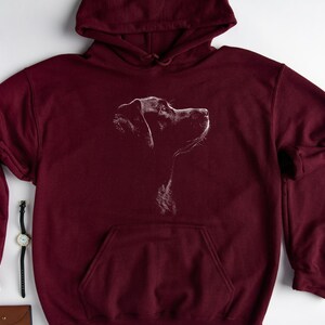 May include: A maroon hooded sweatshirt featuring a white line art graphic of a dog in profile on the chest. The pullover garment includes a drawstring hood, long sleeves with ribbed cuffs, and a large front kangaroo pocket for storage.