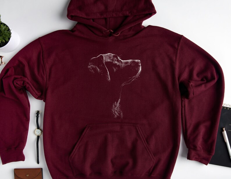 May include: A maroon hooded sweatshirt featuring a white line art graphic of a dog in profile on the chest. The pullover garment includes a drawstring hood, long sleeves with ribbed cuffs, and a large front kangaroo pocket for storage.