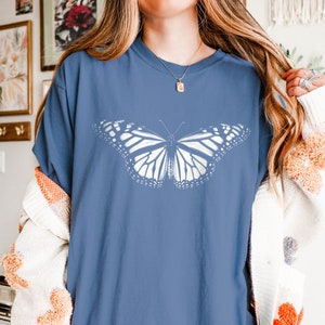 May include: A blue short-sleeved t-shirt featuring a large, white, detailed graphic of a butterfly centered on the chest. The shirt has a crew neckline and a relaxed fit, showcasing the intricate wing patterns of the butterfly in a solid white print.