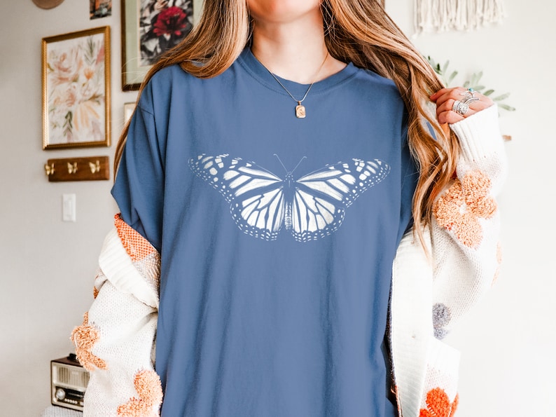 May include: A blue short-sleeved t-shirt featuring a large, white, detailed graphic of a butterfly centered on the chest. The shirt has a crew neckline and a relaxed fit, showcasing the intricate wing patterns of the butterfly in a solid white print.