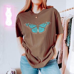 May include: A brown short-sleeve t-shirt featuring a large, centered graphic of a butterfly with bright blue wings and black and white detailing. The shirt has a crew neckline and a relaxed, loose fit, providing a casual look for everyday wear.