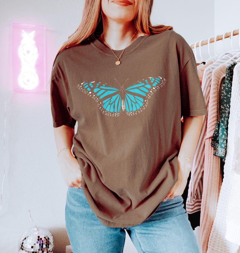 May include: A brown short-sleeve t-shirt featuring a large, centered graphic of a butterfly with bright blue wings and black and white detailing. The shirt has a crew neckline and a relaxed, loose fit, providing a casual look for everyday wear.