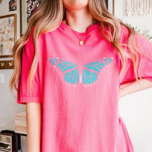 May include: A bright pink short-sleeved t-shirt featuring a large, centered graphic of a butterfly with vibrant turquoise wings and white detailing. The shirt has a relaxed, loose fit with a classic crew neckline and a solid, uniform color throughout.