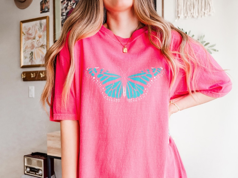 May include: A bright pink short-sleeved t-shirt featuring a large, centered graphic of a butterfly with vibrant turquoise wings and white detailing. The shirt has a relaxed, loose fit with a classic crew neckline and a solid, uniform color throughout.