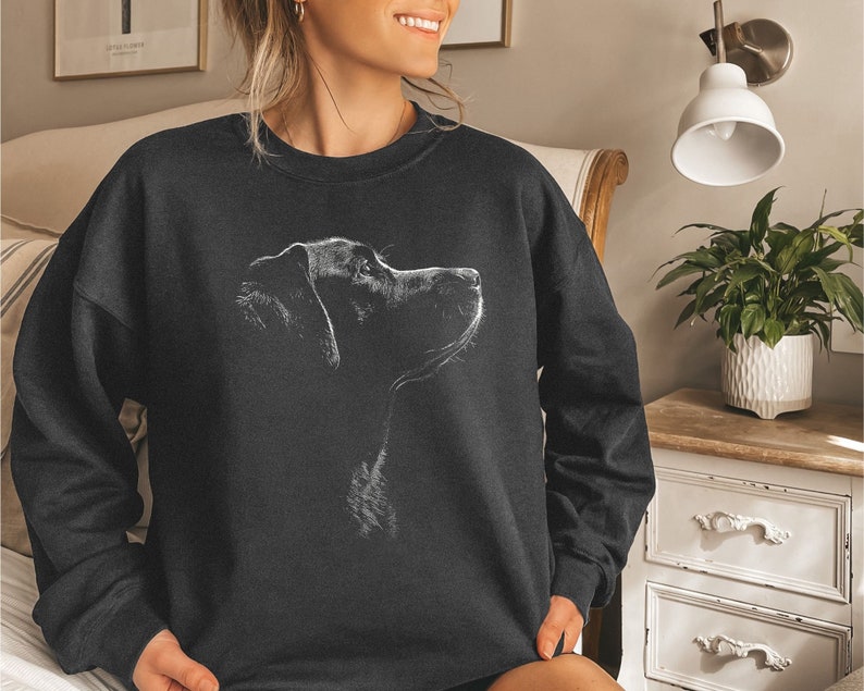 May include: A dark charcoal gray crewneck sweatshirt featuring a white line-art illustration of a dog in profile. The graphic depicts the head and neck of a canine looking upward, rendered in a sketch style against the solid dark fabric background.