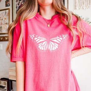 May include: A bright pink short-sleeved t-shirt featuring a large, white, detailed monarch butterfly graphic centered on the chest. The shirt has a crew neckline and a relaxed, loose-fitting silhouette with a solid, vibrant pink color throughout.