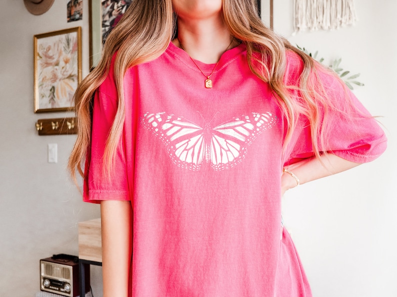 May include: A bright pink short-sleeved t-shirt featuring a large, white, detailed monarch butterfly graphic centered on the chest. The shirt has a crew neckline and a relaxed, loose-fitting silhouette with a solid, vibrant pink color throughout.