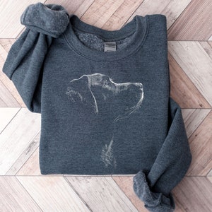 May include: A dark heather gray crewneck sweatshirt featuring a white line art illustration of a dog in profile. The design depicts the head and neck of the canine facing toward the right side, rendered in a sketch-like style on the front center.