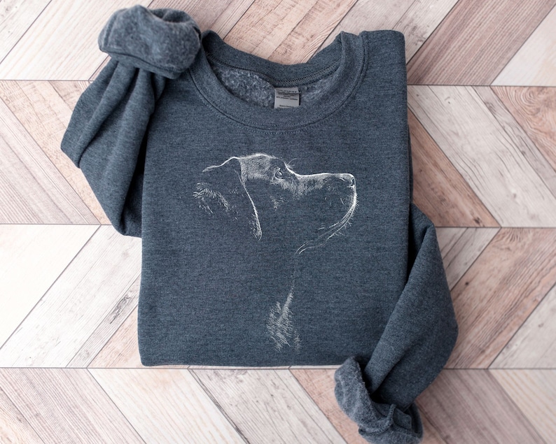 May include: A dark heather gray crewneck sweatshirt featuring a white line art illustration of a dog in profile. The design depicts the head and neck of the canine facing toward the right side, rendered in a sketch-like style on the front center.