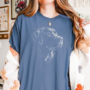 Op de afbeelding: Een blauw t-shirt met korte mouwen en een witte lijntekening van een hondenkop in profiel op de borst. Het shirt heeft een ronde hals en een relaxte pasvorm, met een gedetailleerde schets van een hondenhoofd met hangende oren.