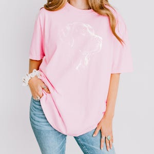Op de afbeelding: Een lichtroze T-shirt met korte mouwen en een witte lijntekening van een hondenkop op de borst, gecombineerd met een blauwe spijkerbroek. Het shirt heeft een ronde hals en een relaxte pasvorm met een eenvoudig grafisch ontwerp.