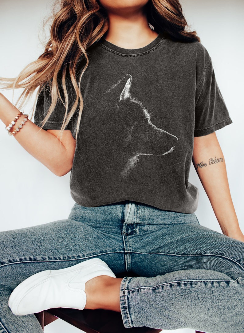May include: A charcoal grey short-sleeve t-shirt featuring a white line-art graphic of a dog's profile on the chest. The shirt has a crew neckline and a faded, vintage-style wash. The design depicts the head and ears of a canine in silhouette.