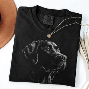 Op de afbeelding: Een zwart T-shirt met korte mouwen en een witte lijntekening van een hondenkop op de voorzijde. Het label in de nek bevat de tekst Comfort Colors, S-P-CH en 100% cotton. Het T-shirt is gemaakt van een effen zwarte stof.